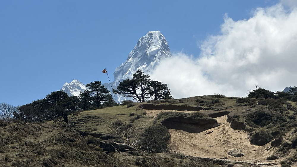 A dusty, grassy hill is in the foreground. A huge, snow-capped mountain peak protrudes from behind.