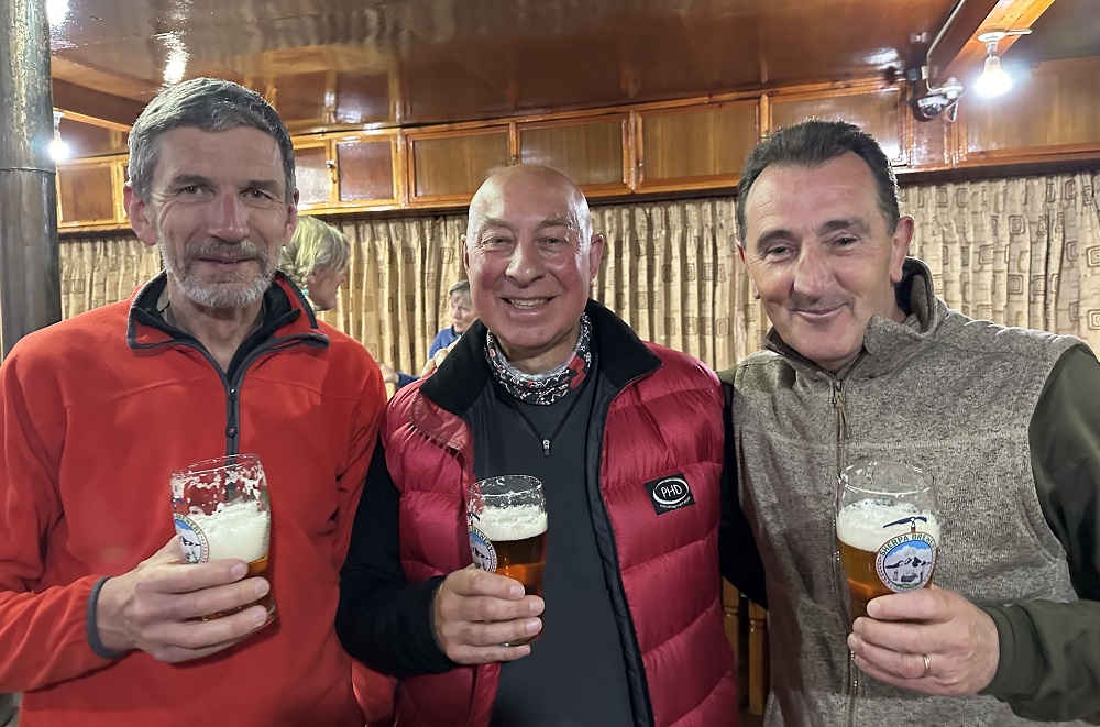 The three men stand smiling in fleeces inside a room with a wooden ceiling. Each of them is holding a pint of beer.