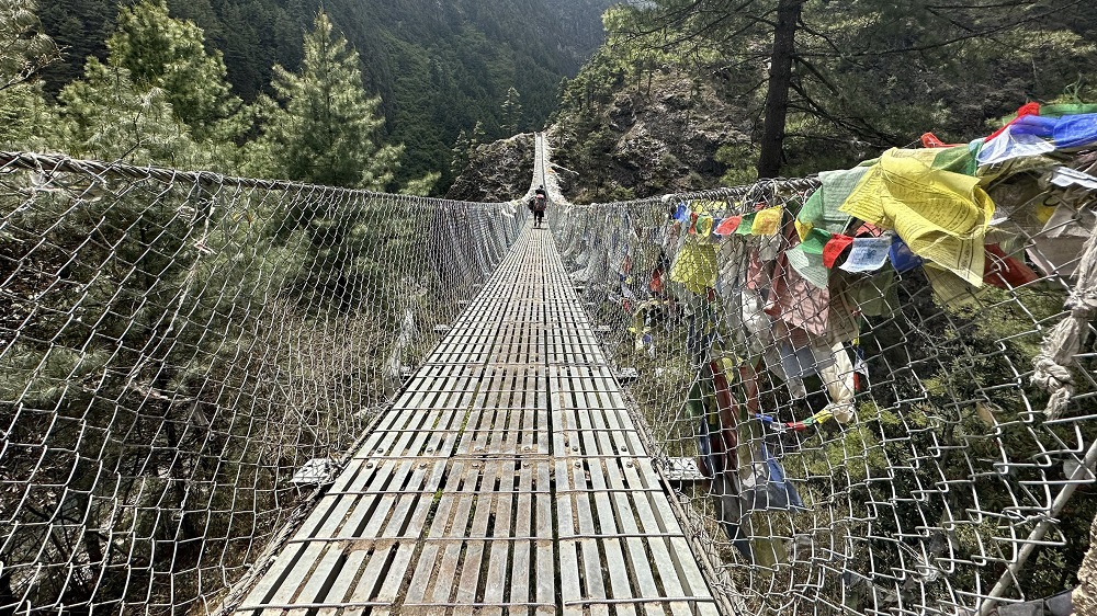 A wire bridge with wooden slats hands over a river valley. Coloured flags are tied to the sides.