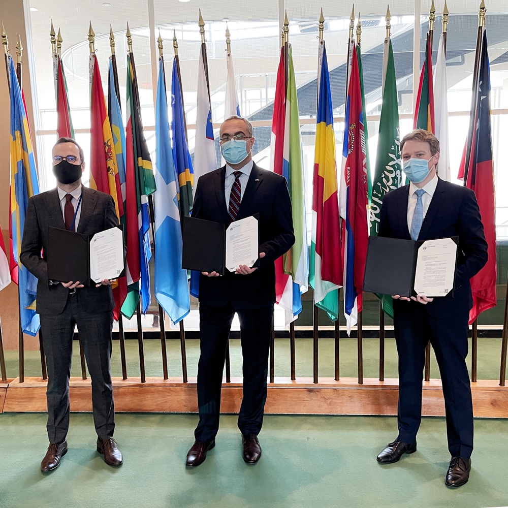 Robin and the two other representatives stand holding documents open facing the camera. They are each dressed in black suits and wearing face masks. Behind them are numerous colourful flags on poles.