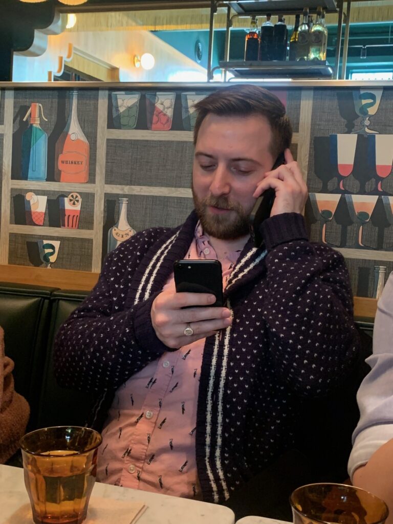 Britton speaks on one phone while holding another. He is sat in casual clothes at a restaurant with a glass of water in front of him.