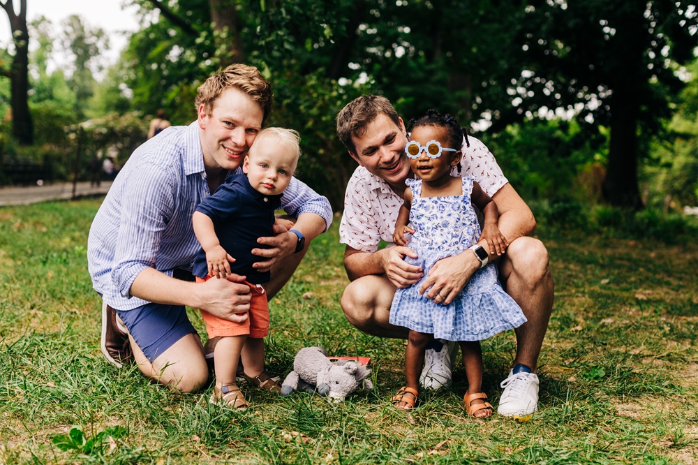 Robin and his partner both crouch down. Each has a small, standing child in front of them. Robin has his arms around Remi, a little boy with blond hair, a blue shirt and orange shorts. Matt is holding Louise who has black braided hair, a blue sundress and flower sunglasses.