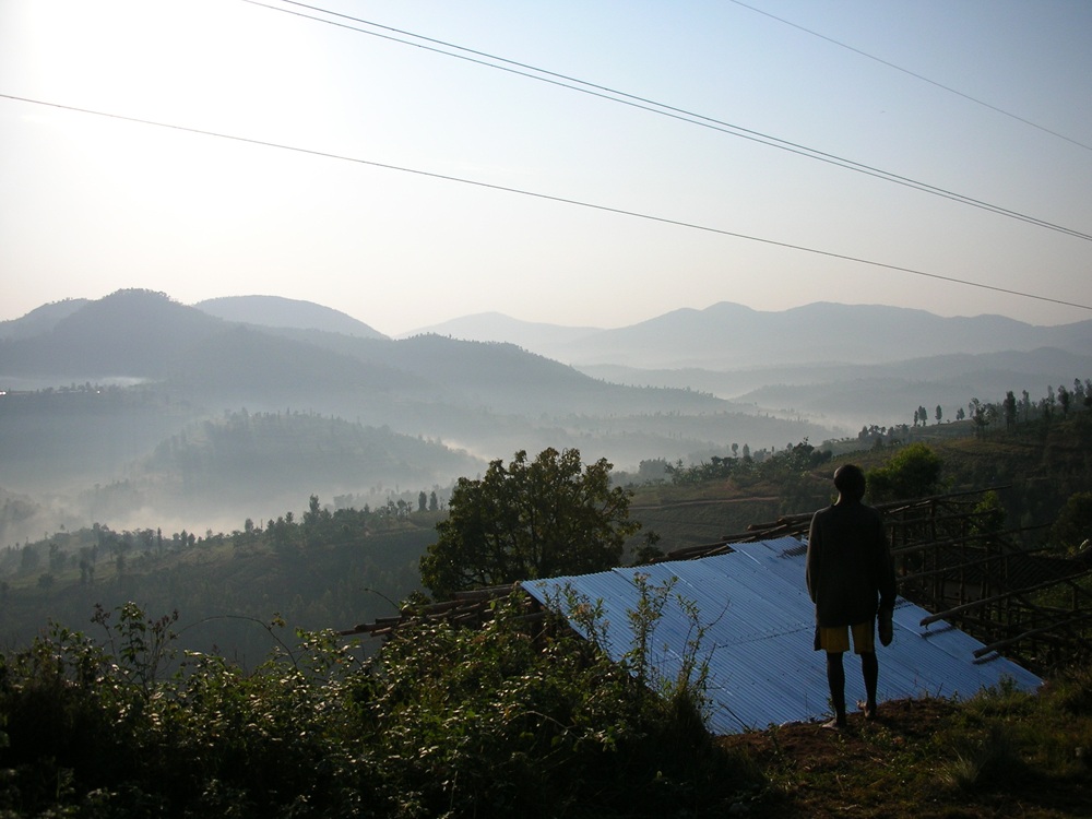 A man stands in front of a roof on a hill in Rwanda. In the sky are powerlines and in the distance are mist-covered hills.