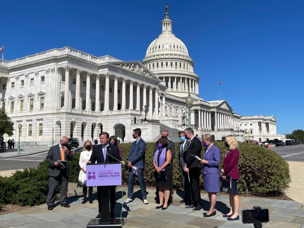 Members of congress gather around a speaker with a podium. The podium reads 'Bipartisan addiction and mental health taskforce'.