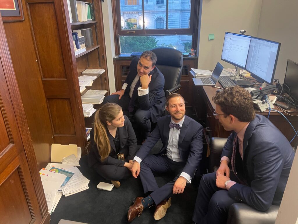 Britton sits on the floor in a messy office. Three colleagues sit around him, one on the floor and two on chairs. They are all wearing smart suits.