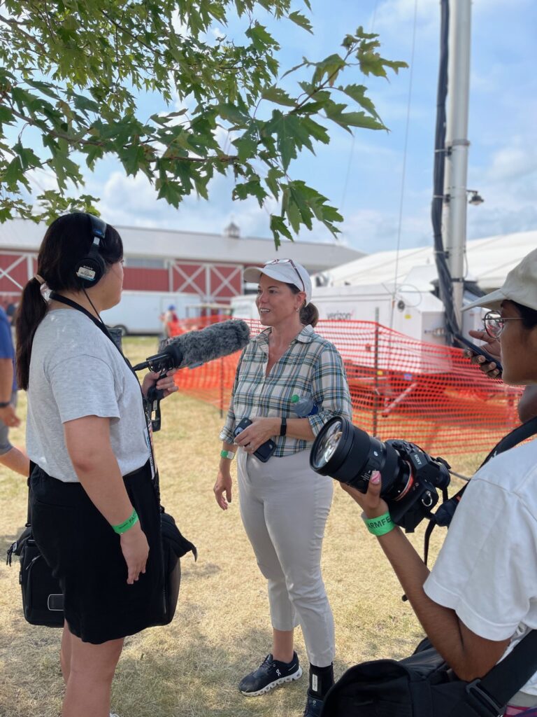 One person in a white baseball cap talks to an interviewer with a sound pole and headphones. Another person stands to the side with a camera.