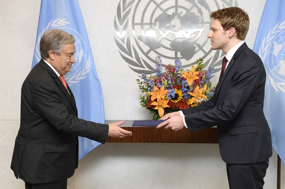 Antonio Guterres stands on the left facing Robin. Robin holds a blue book towards him. Behind them are the blue UN flags, a metal wall relief of the UN logo and a nice display of colourful flowers.