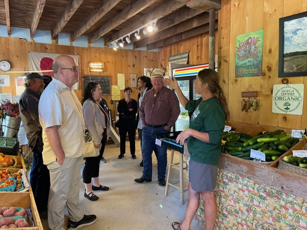 People in casual clothes gather in around a speaker in a green sweatshirt in a farm shop. The walls are wood panelled and fresh produce sits out in wooden crates.