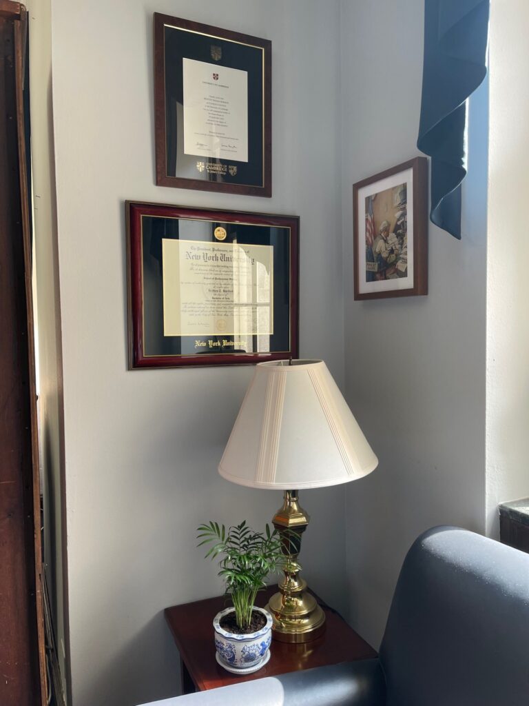 A lamp and a small pot plant sit on a brown side table. Above them on the wall Britton's SJC diploma and another certificate and a photo are hung.