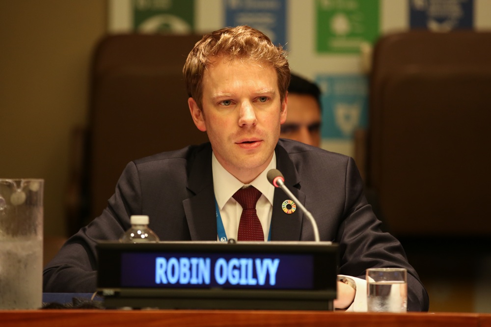 Robin sits with a microphone and his name on an electronic sign in front of him.