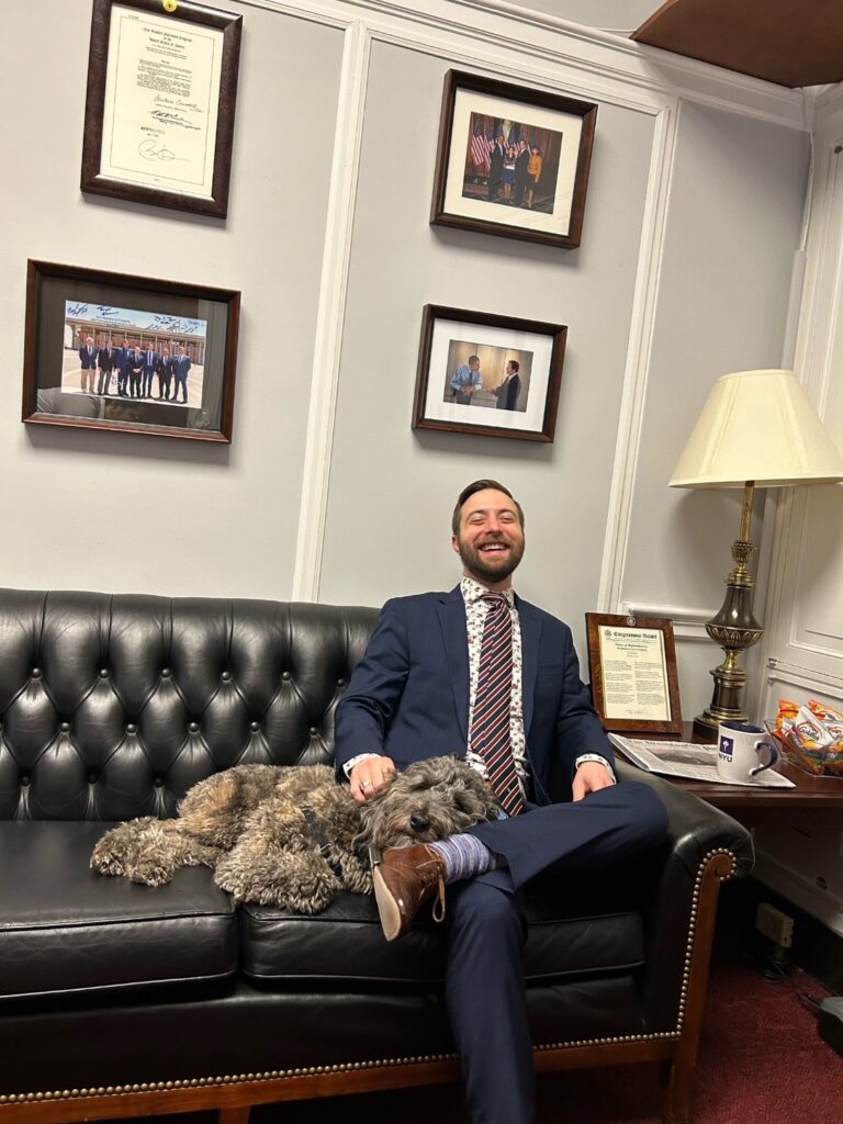 Britton sits on a couch wearing a dark blue suit and stripy tie. A fluffy grey dog lies across his lap. One the walls are framed photos and certificates.