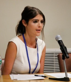 Alexandra is wearing a white sleeveless shirt and a lanyard. She is speaking into a microphone.