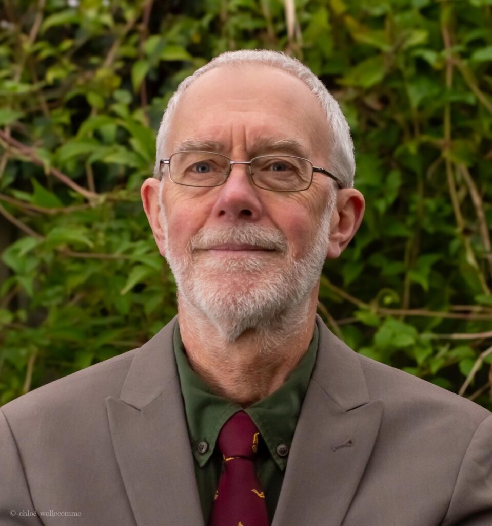 A head and shoulders image of Paul. He is wearing a grey suit, green shirt and red tie. He has short grey hair and glasses.