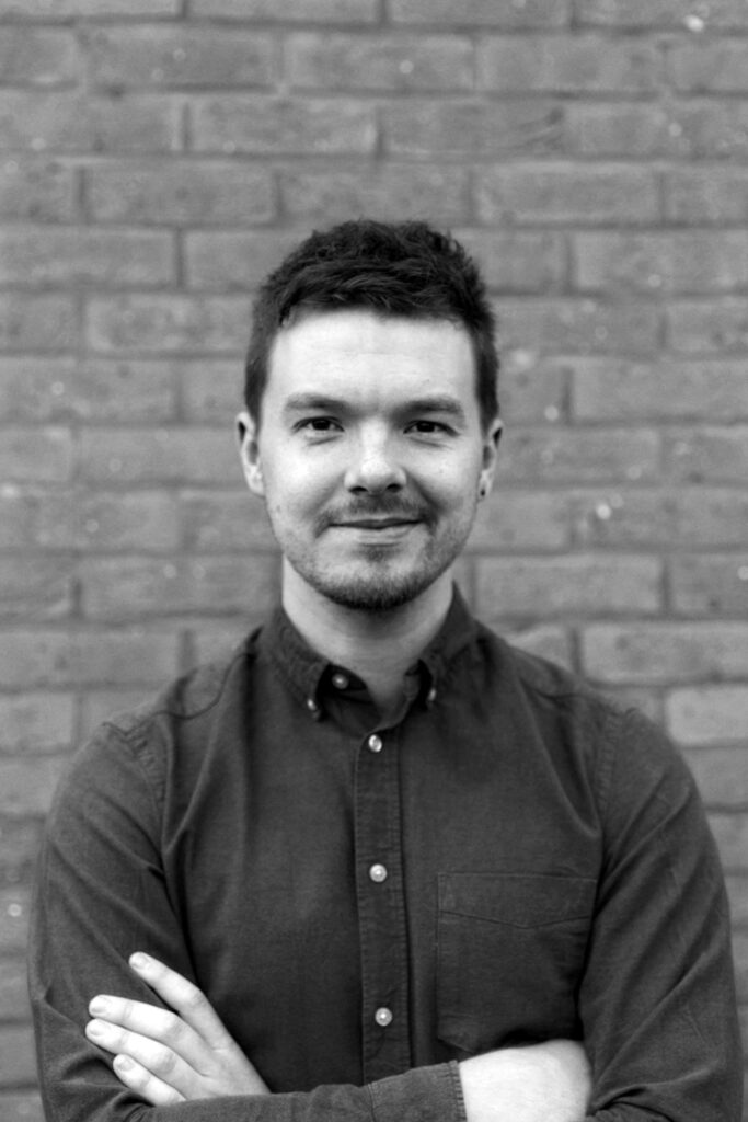 A black and white headshot of Simon. He is wearing a dark buttoned shirt, with his arms folded. He is smiling.