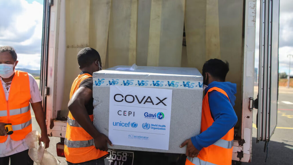 Two men in high-vis vests carry a large crate of vaccines. The box reads 'COVAX' in large letters.