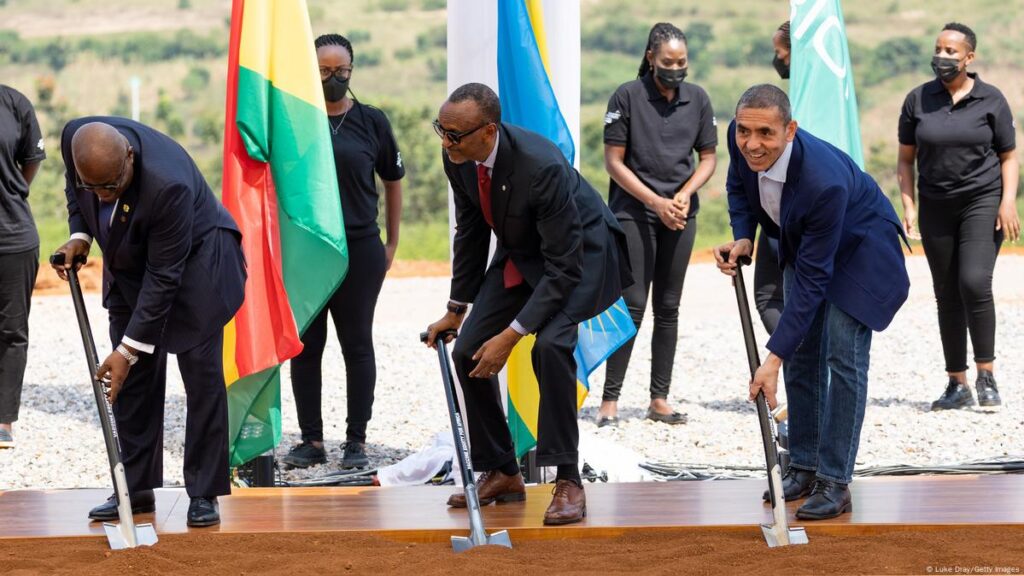 Three men in suits ceremonially dig the ground with shovels. Behind them are some colourful flags and women in face masks.