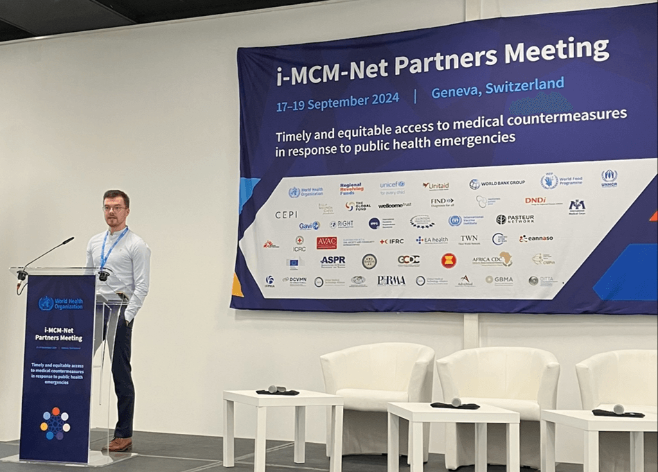 Simon stands at a podium speaking. Beside him is a large poster which reads: 'i-MCM-Net Partners Meeting' and then 'Timely and equitable access to medical countermeasures in response to public health emergencies'.