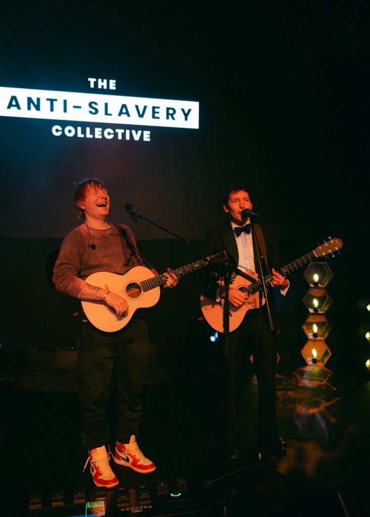 Ed Sheeran and James Blunt hold guitars on stage and sing into microphones.